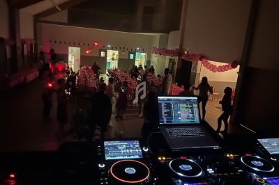 Vue depuis une table de mixage sur une salle décorée avec des ballons roses, des invités dansent et discutent.