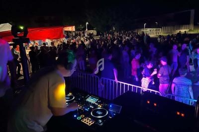Un DJ mixe sur scène en plein air devant une foule dansante sous des lumières colorées.