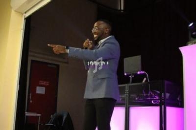 Un homme en costume parle au micro sur une scène éclairée, avec une table de mixage en arrière-plan.