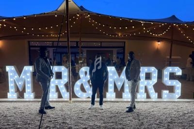 Trois personnes posent devant une enseigne lumineuse ’MR & MRS’ sous un ciel crépusculaire avec des guirlandes suspendues.