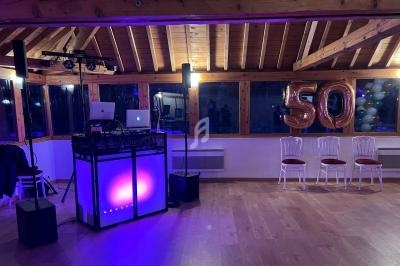Salle décorée pour une fête, avec une table de DJ éclairée, ballons ’50’ et chaises alignées près des fenêtres.