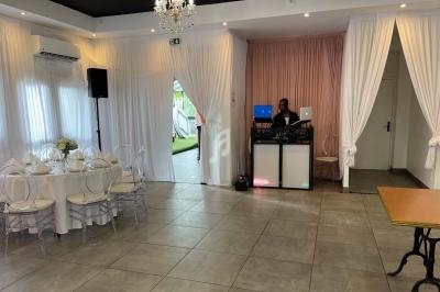 Salle de réception lumineuse avec table dressée, rideaux blancs et DJ installé près d'un mur rose.