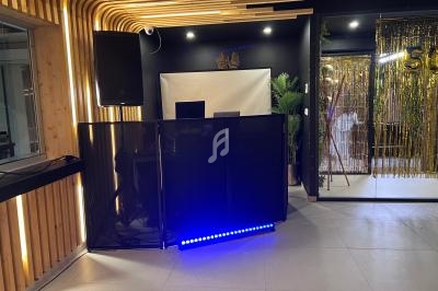 Pupitre de DJ noir avec éclairage LED bleu, installé dans une salle moderne décorée de bois et de rideaux dorés.