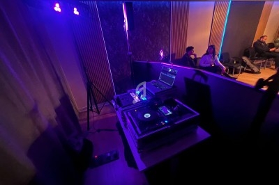 Équipement de DJ avec platines et ordinateur dans une salle faiblement éclairée, quelques personnes assises en arrière-plan.
