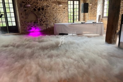 Salle en pierre avec sol en carrelage, éclairage coloré et fumée dense couvrant le sol devant une table drapée.