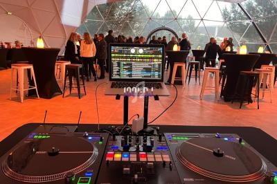 Poste de DJ avec platines et ordinateur portable, installé dans une salle vitrée où des invités discutent debout autour de…