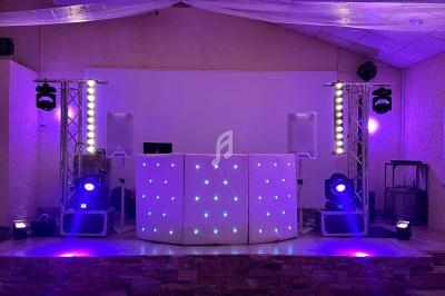 Scène de soirée avec une table de DJ illuminée, projecteurs colorés et enceintes dans une salle au plafond en poutres…