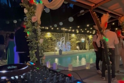 Console de DJ en premier plan avec des invités dansant sous une pergola décorée de lumières et de fleurs.