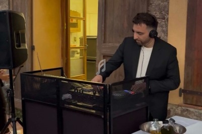 Un DJ en costume mixe de la musique derrière une table équipée, dans un espace intérieur avec des murs en pierre.