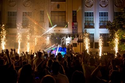 Foule en fête devant une scène illuminée par des feux d'artifice et des lasers, devant un bâtiment Art déco.