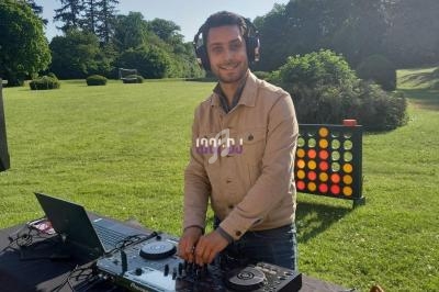 Un DJ mixe en plein air sur une table équipée de platines, avec un grand jardin verdoyant en arrière-plan.