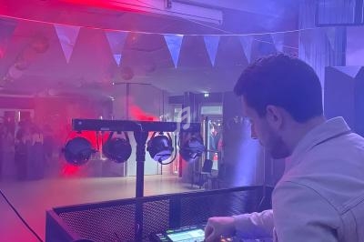 Un DJ mixe sur une console dans une salle éclairée par des lumières rouges et violettes, avec des invités en arrière-plan.