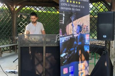 Un DJ mixe sous un kiosque en bois, avec une bannière promotionnelle et du matériel audio installé devant lui.