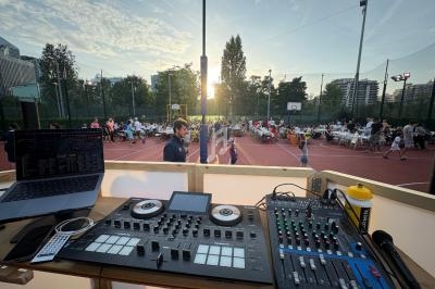 Vue d'une table de mixage et d'un ordinateur surplombant un terrain de sport avec des tables et des convives en plein air.