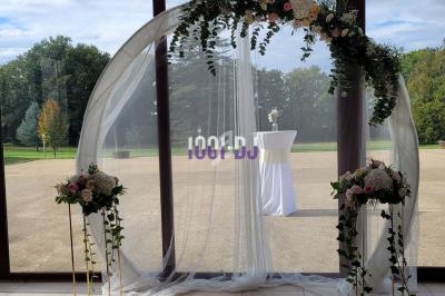 Arche de mariage circulaire décorée de fleurs et de voilages blancs, placée devant une baie vitrée avec vue sur un jardin.