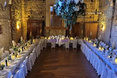 Salle en pierre avec deux longues tables dressées pour un banquet, décorée de fleurs suspendues et éclairée par des bougies.