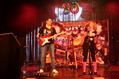 Deux musiciens sur scène jouent de la guitare et chantent devant une décoration sur le thème des années 80.