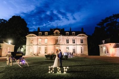 Un couple se tient devant un château éclairé la nuit, avec des initiales lumineuses ’M & F’ sur la pelouse.