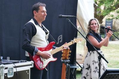 Un homme joue de la guitare électrique rouge et une femme chante devant des micros en extérieur.