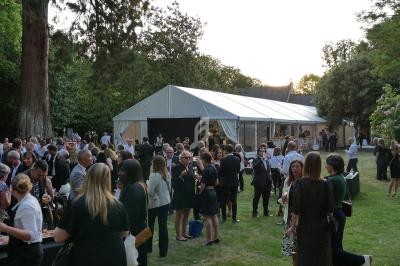 Groupe de personnes rassemblées dans un jardin lors d'un événement en plein air avec une grande tente blanche en arrière…