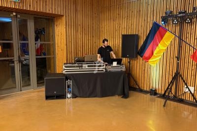 Un DJ mixe derrière une table équipée, entourée de drapeaux français et allemand dans une salle en bois.