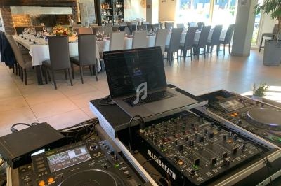 Salle de réception lumineuse avec tables dressées, vue depuis une table de mixage et des platines DJ au premier plan.