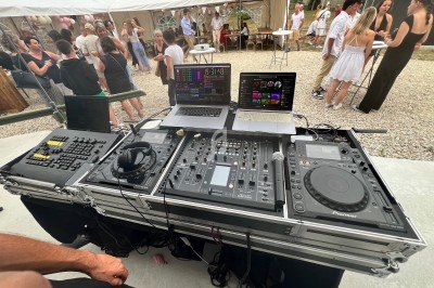Poste de DJ avec table de mixage, platines, ordinateurs et contrôleurs, lors d'un événement en plein air avec des invités.