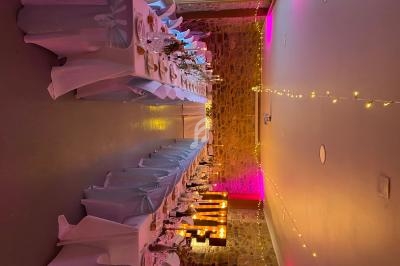 Salle décorée pour un événement avec longues tables dressées, nappes blanches, guirlandes lumineuses et mur en pierre.