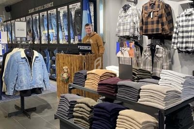 Un magasin de vêtements avec des jeans accrochés au mur, des piles de pulls et chemises, et un vendeur souriant au comptoir.