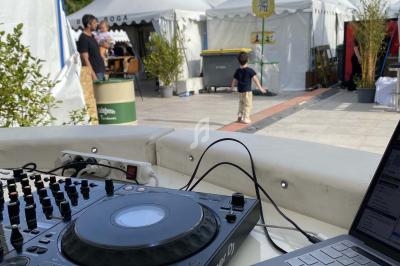 Vue d'une table de mixage et d'un ordinateur portable en extérieur, avec des stands et des personnes en arrière-plan.