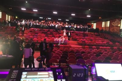 Salle de spectacle avec sièges rouges, public debout en arrière-plan et matériel technique visible au premier plan.