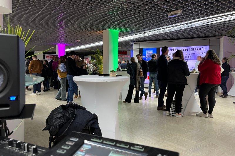Des personnes discutent autour de tables hautes dans un espace éclairé par des lumières colorées, avec un stand en arrière…