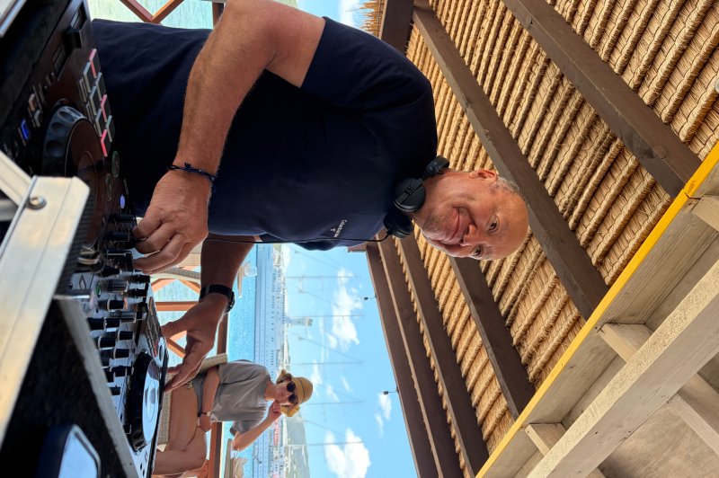 Un DJ mixe de la musique en extérieur sous une structure en bois, avec vue sur la mer et un port en arrière-plan.