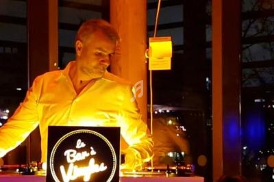 Un homme en chemise claire se tient derrière une table lumineuse avec un panneau ’Bar à Vinyles’ dans un espace sombre.