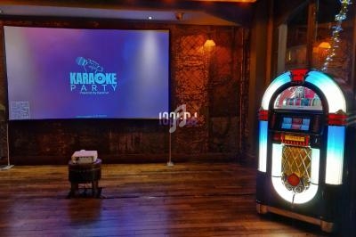 Salle de karaoké avec un jukebox lumineux à droite et un écran de projection affichant ’Karaoke Party’.