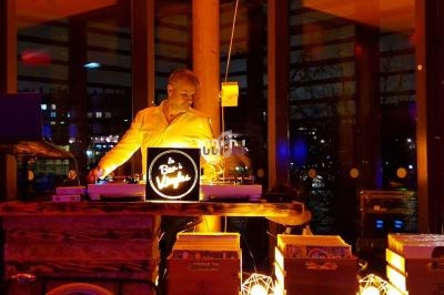 Un DJ mixe sur un vélo aménagé en stand lumineux, dans un espace vitré avec vue nocturne sur une rivière.