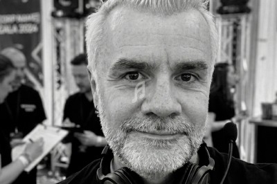 Homme souriant avec un casque audio autour du cou, dans un environnement d'événement avec des lumières et des personnes en…