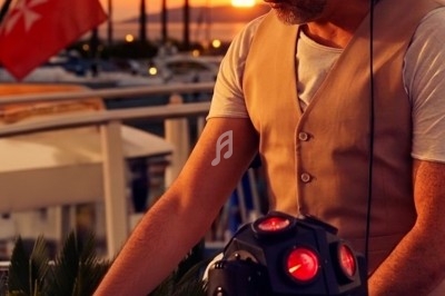 Un DJ mixe en plein air au coucher du soleil, avec des palmiers et un drapeau en arrière-plan.