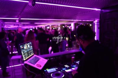 Un DJ mixe de la musique sur une console dans une salle éclairée par des lumières violettes, avec des personnes en arrière…