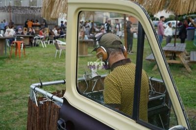 Un DJ mixe en plein air depuis un véhicule aménagé, avec des tables et des personnes en arrière-plan.