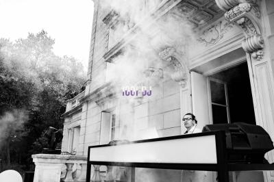 Un DJ mixe en extérieur devant un bâtiment ancien, entouré de fumée et d'équipements audio.