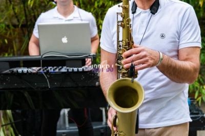 Un musicien joue du saxophone au premier plan, accompagné d'un DJ mixant en arrière-plan dans un cadre extérieur.