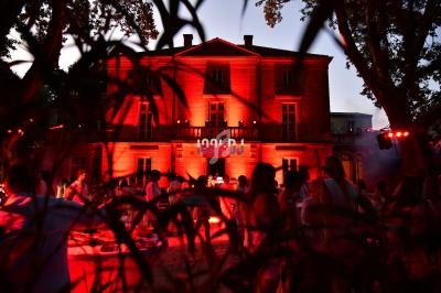 Façade d'un bâtiment illuminé en rouge au crépuscule, avec une foule en contrebas et des branches au premier plan.