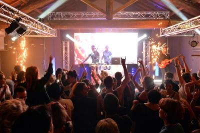 Foule dansant devant une scène avec DJ, éclairages colorés, effets pyrotechniques et ambiance festive en intérieur.