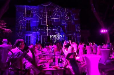 Des invités en tenue blanche assistent à un événement nocturne avec des projections lumineuses sur un bâtiment.