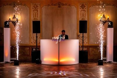 Un DJ mixe derrière une table lumineuse entourée de colonnes lumineuses et d'effets pyrotechniques dans une salle élégante.