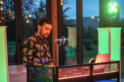 Un DJ mixe de la musique sur une console éclairée, dans une salle vitrée avec vue sur l'extérieur au crépuscule.