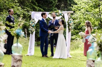Un couple échange ses vœux devant un officiant sous une arche décorée, entouré de témoins dans un jardin verdoyant.