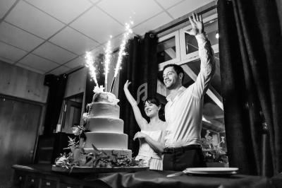 Un couple souriant près d'un gâteau de mariage décoré, avec des cierges magiques allumés, dans une salle festive.