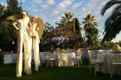 Deux artistes sur échasses vêtus de blanc dans un jardin, entourés de tables dressées pour un événement en plein air.